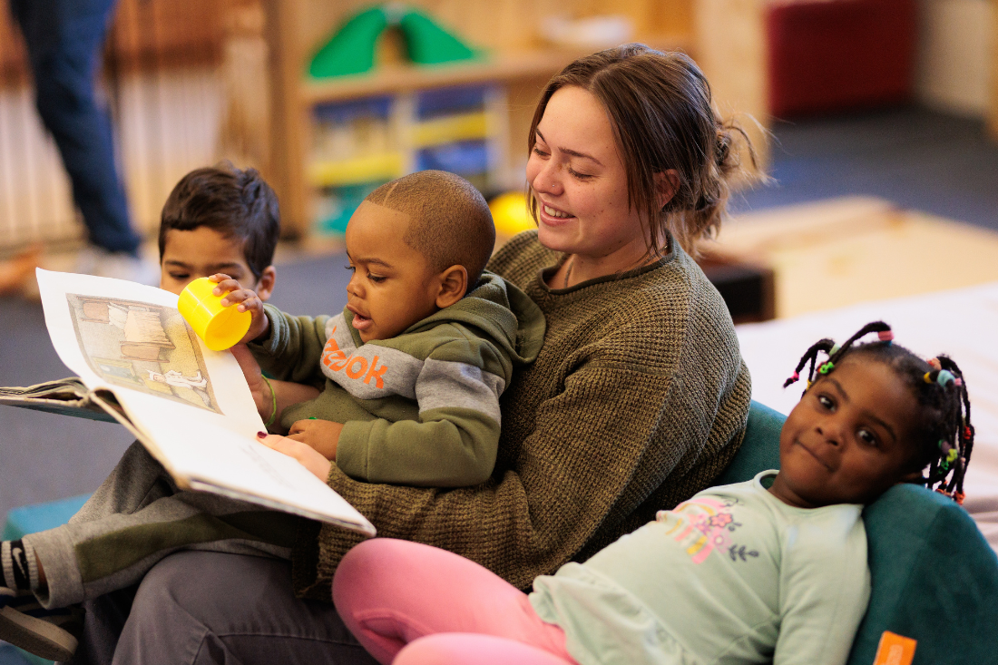 An Early Childhood Educator reads a book to three young children.